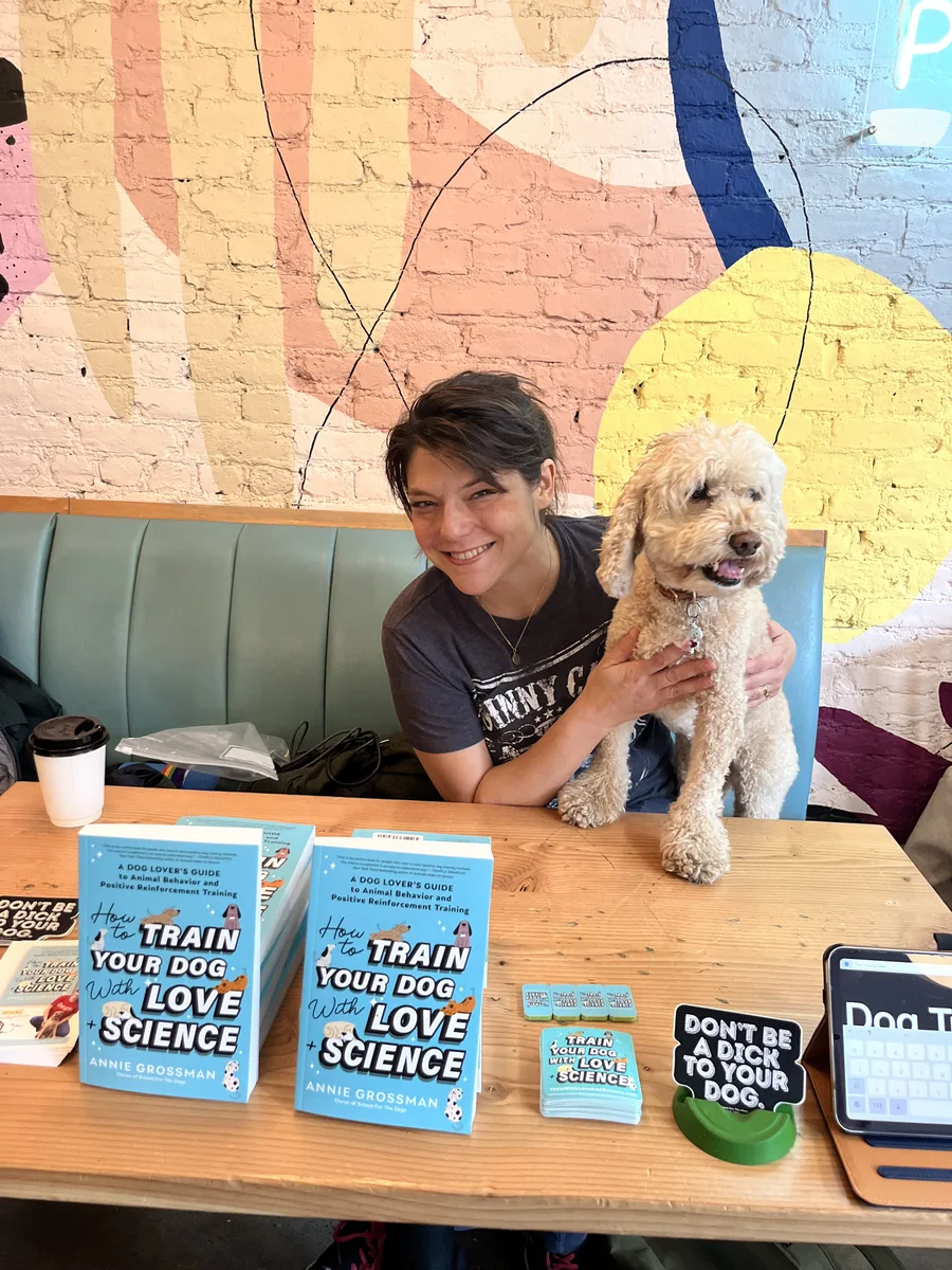 Annie Grossman with a doodle at a book signing table stacked with How To Train Your Dog With Love + Science